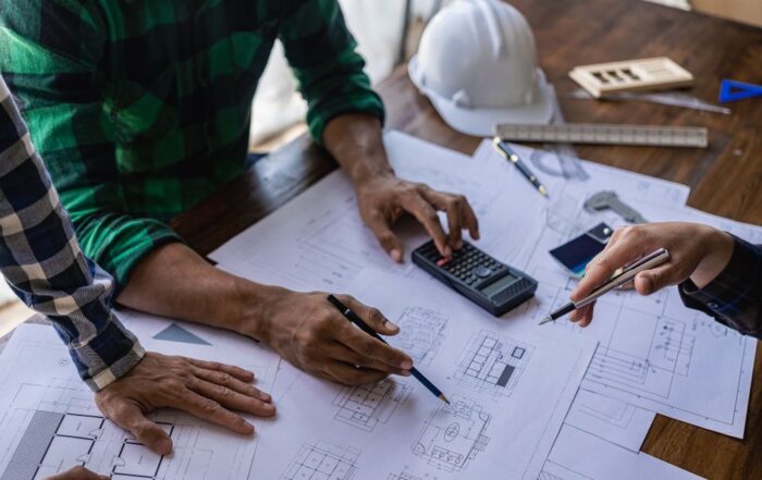 A group of professionals sits over a table reviewing blueprints with a compass, hardhat and more construction-based items, signifying KIP large format printing.