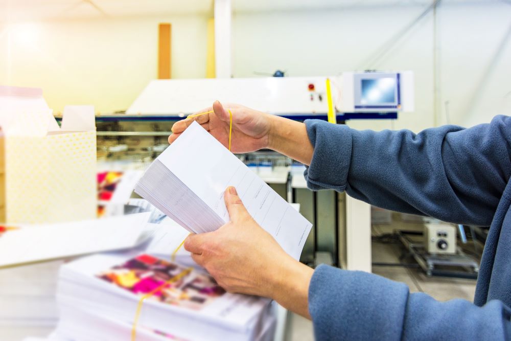 A worker's hands folding sheets of paper freshly printed from a digital press in a busy print shop environment.