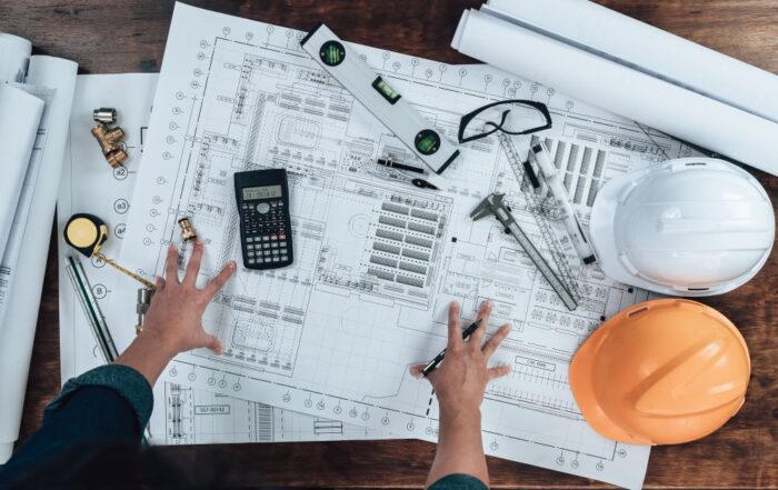 An engineer or architect reviewing blueprints on a desk filled with tools, hard hats, and measuring instruments. The detailed plans highlight the importance of using a wide format printer for producing accurate and high-quality large-scale documents needed for construction and design projects.